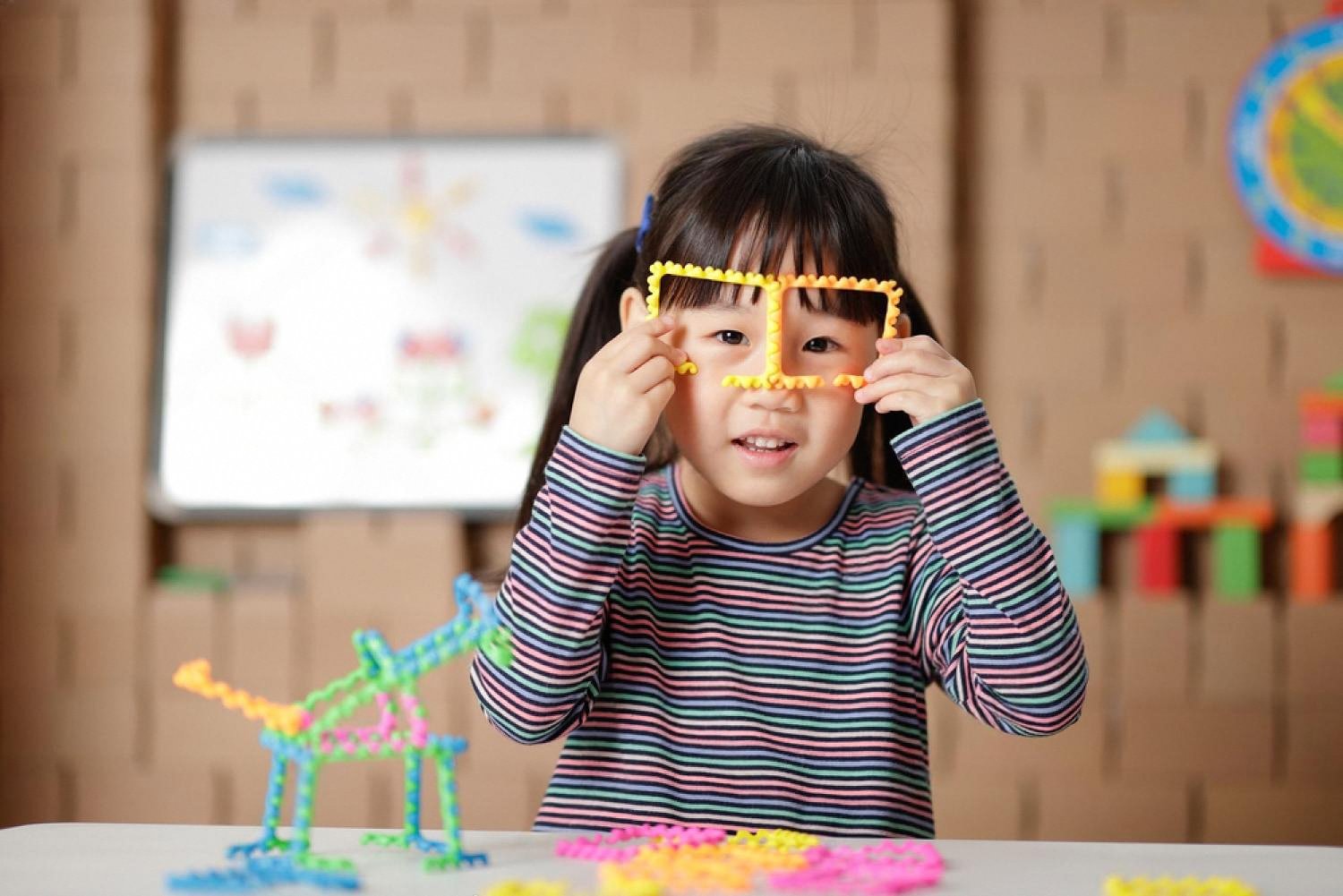 Girl building a colourful 3D figure from craft pieces at a table with toys behind her